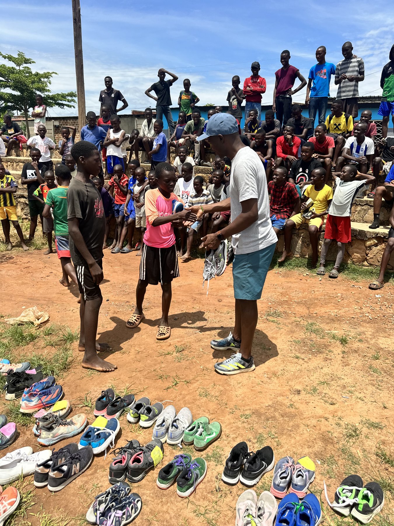 Prince Mumba handing a pair of running shoes to a boy at a shoe distribution in Zambia, with children watching